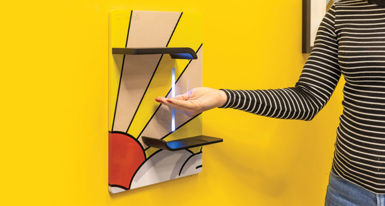 Woman in black and white striped shirt holds hand under Vaask faucet to receive hand sanitizer for unit with a sunrise custom graphic displayed on it.